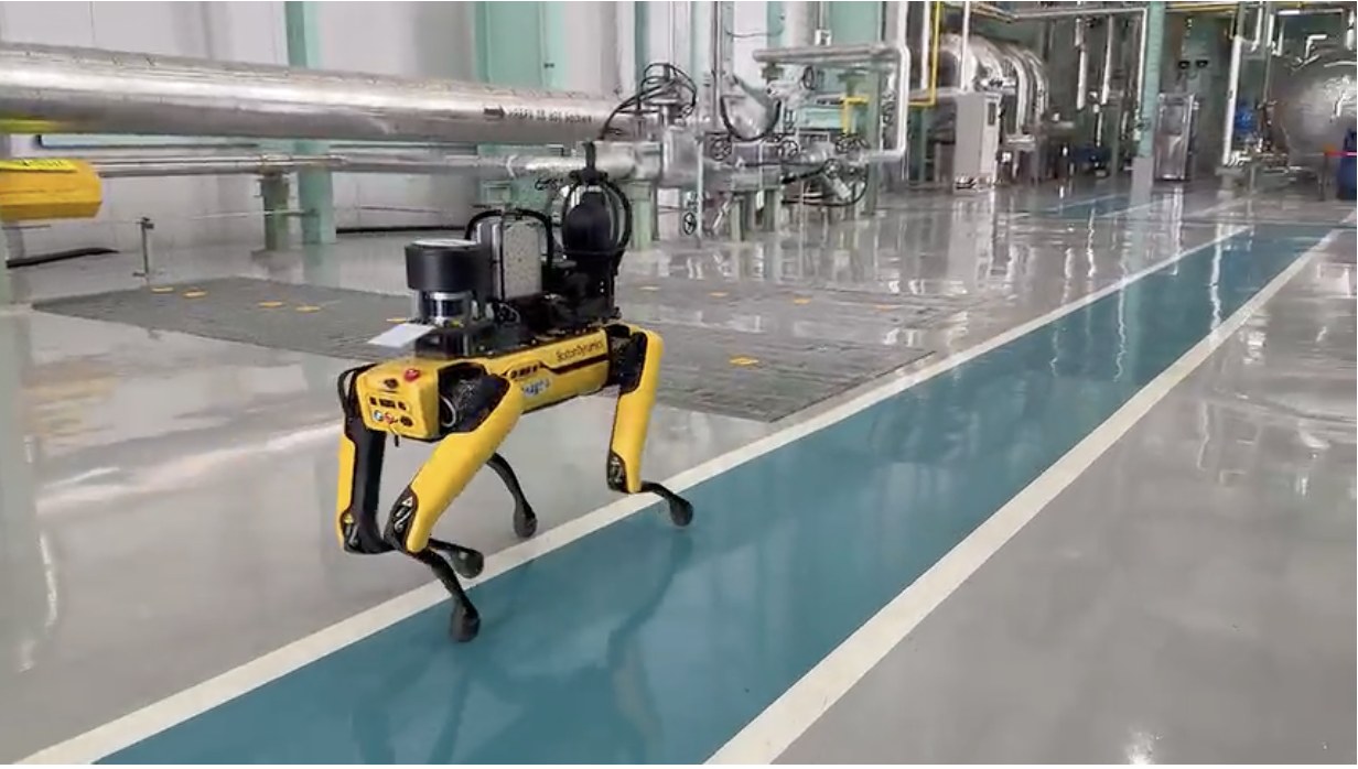 A yellow Boston Dynamics Spot robot carrying a sensor payload walks a painted aisle past pipes and valves inside a power plant