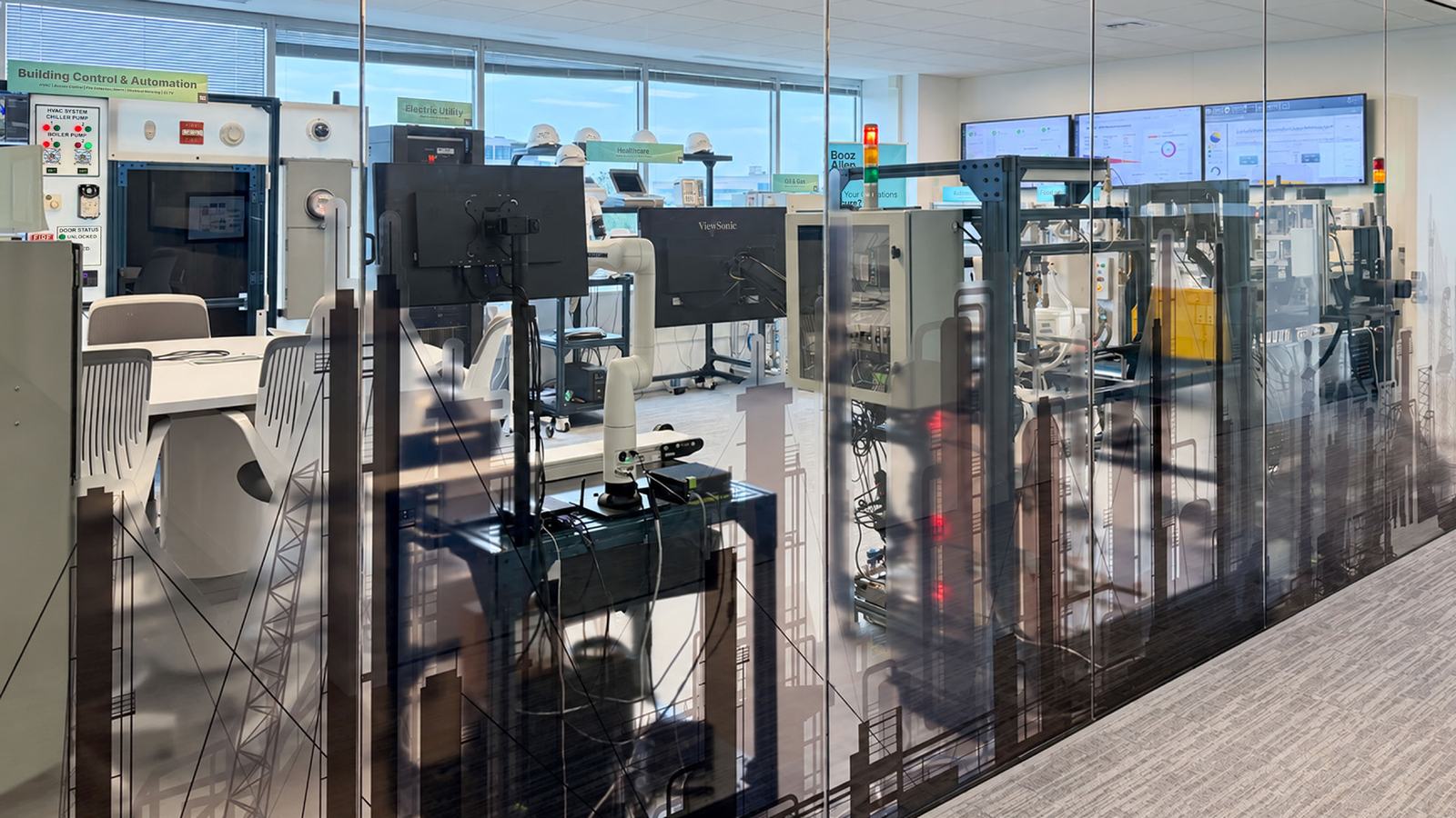 Booz Allen's operational technology lab seen through a glass wall, with a building control and automation panel, a robotic arm on a test bench, equipment racks and monitoring screens