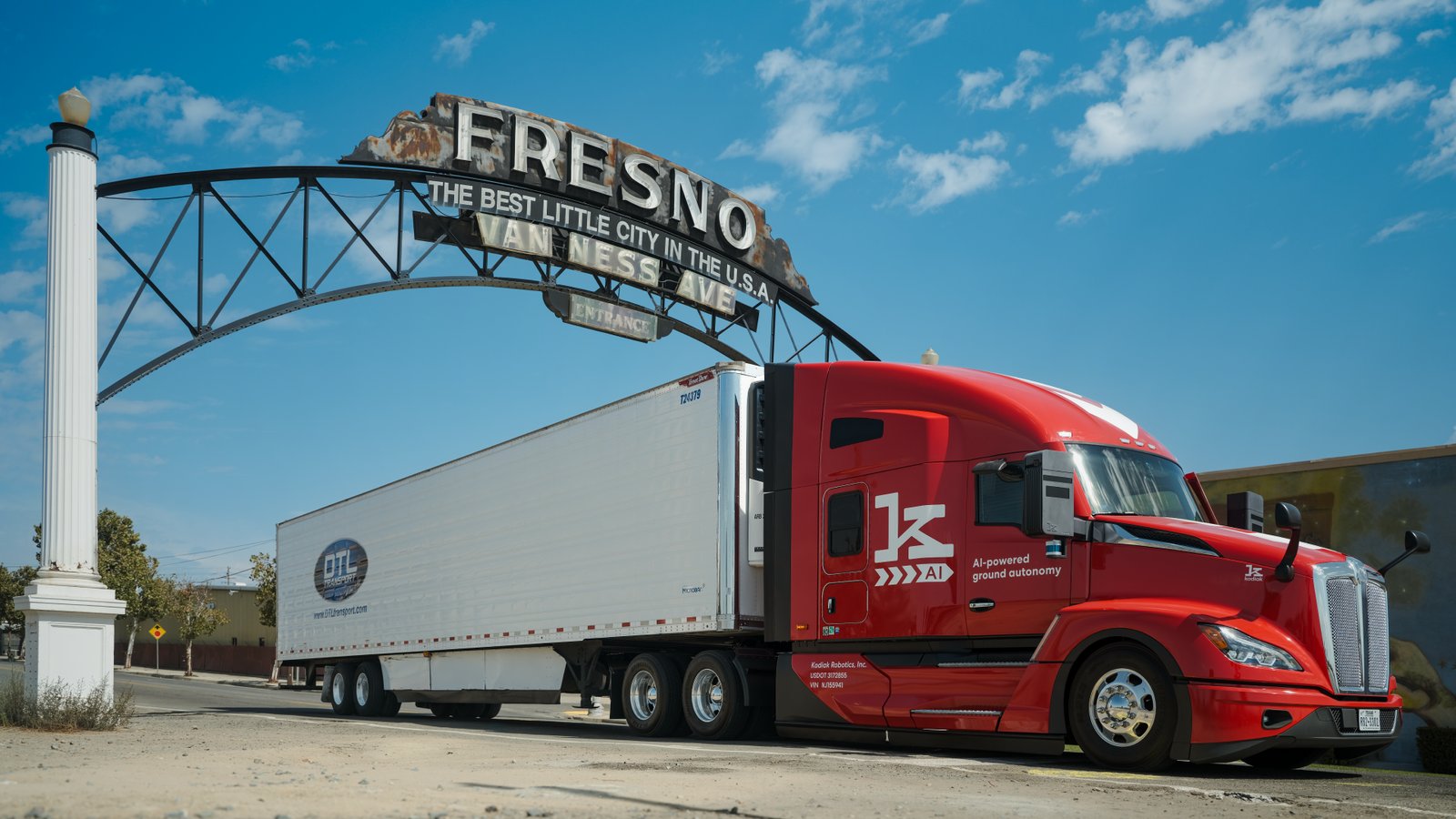 A red Kodiak AI-equipped semi truck pulling a white DTL trailer parked beneath the historic Fresno Van Ness Avenue arch sign under a blue sky