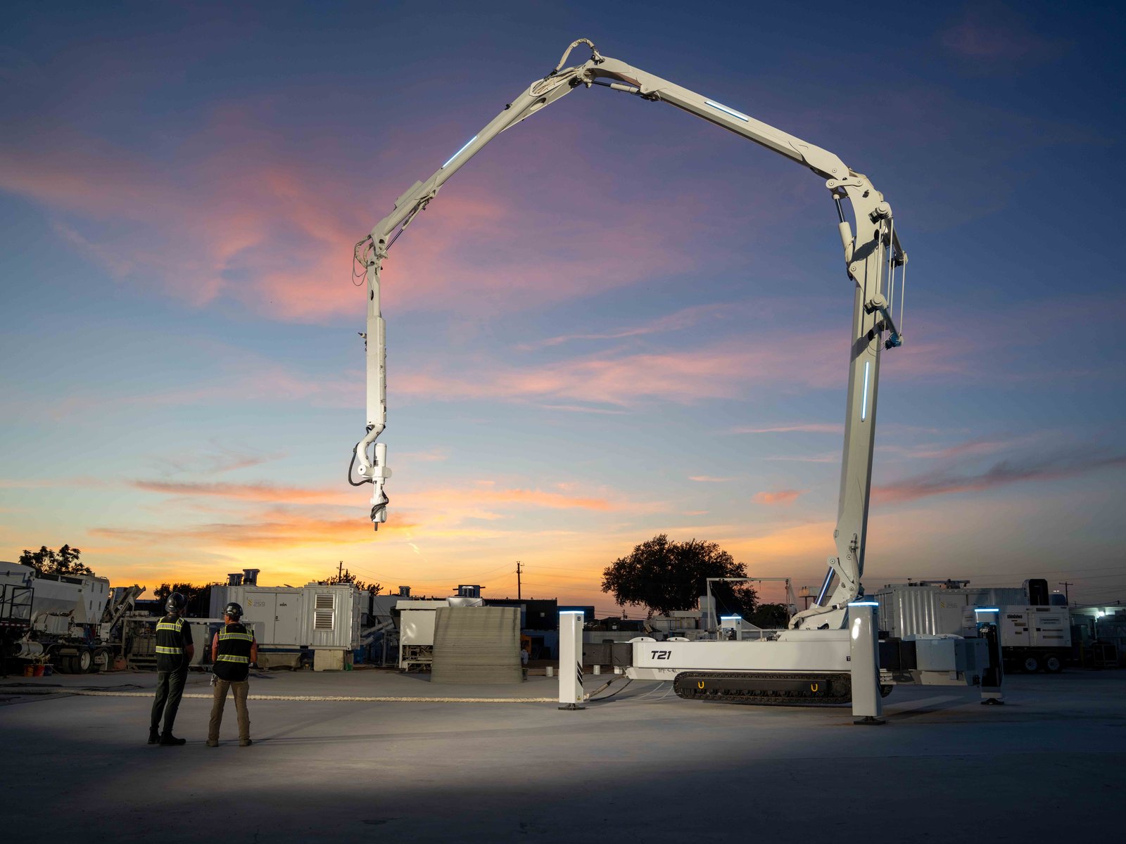 ICON's Titan printer, a white tracked base with a long boom arm, at its Austin R&D facility at dusk with two workers in safety vests