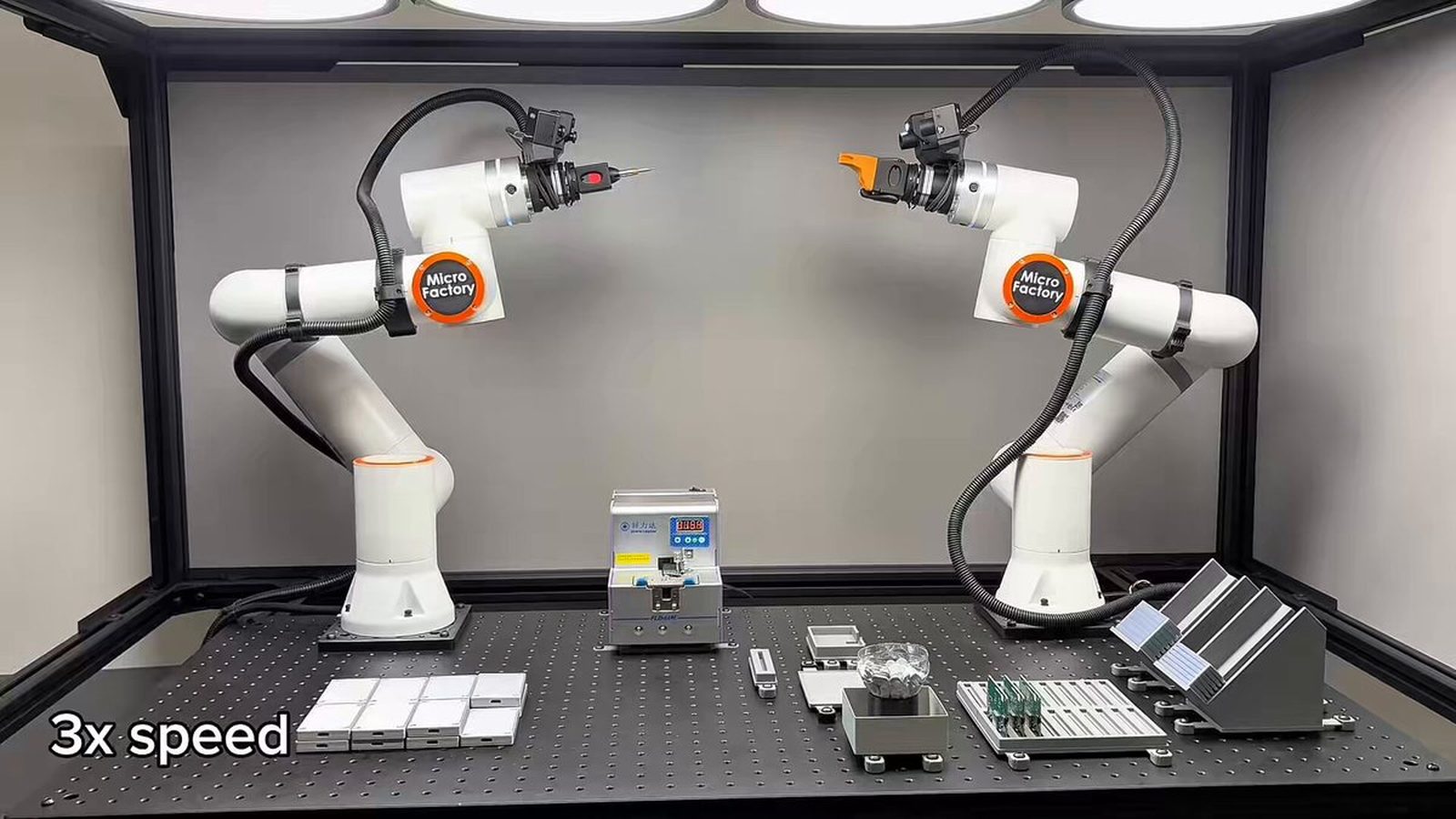 Two white MicroFactory robot arms facing each other inside an enclosed workcell, one holding a screwdriver bit and one an orange gripper, above a perforated table with a screw feeder, stacked white parts, a parts tray and a circuit board fixture, with a "3x speed" label from the video