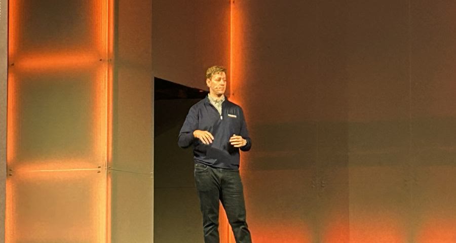 A man in a dark suit jacket speaking into a headset microphone on a conference stage